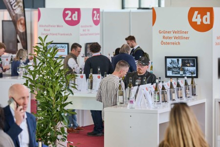 Visitors taste wines and non-alcoholic wines at a trade fair stand with clear labeling according to variety and origin.